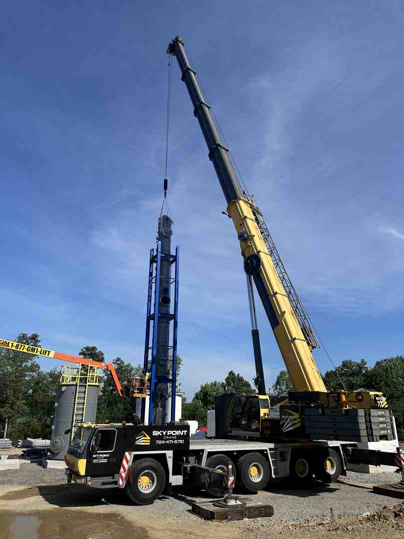Crane lifts a tall blue column-mounted structure on a construction site with a yellow crane nearby under a clear blue sky.