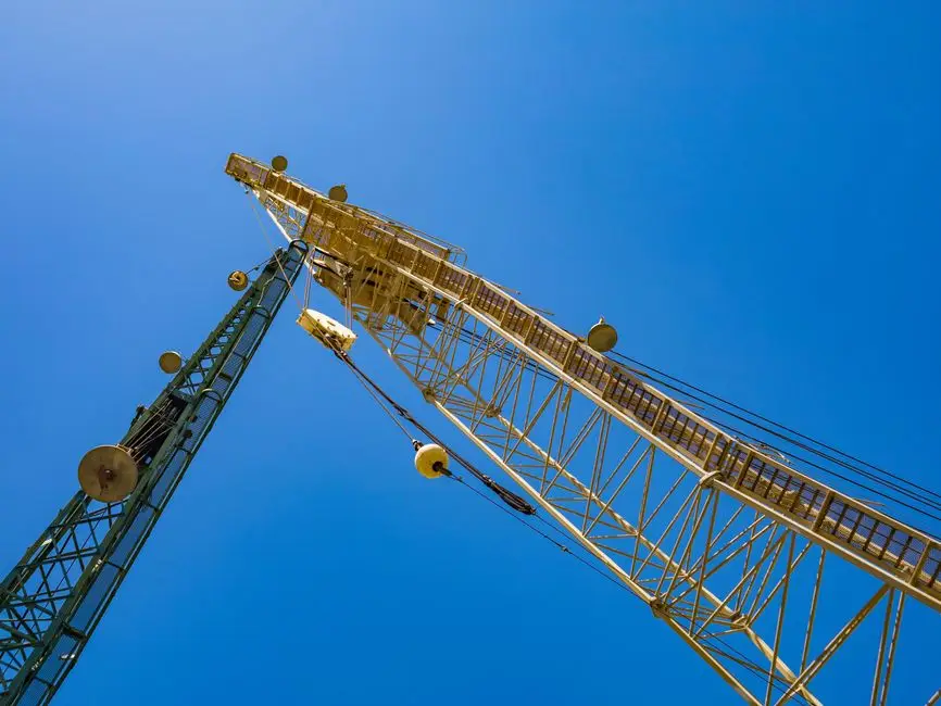 Tall construction crane reaching into a clear blue sky against metal framework and cables.